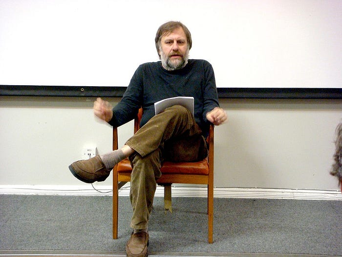 Slavoj Zizek sitting on a wooden chair in front of a whiteboard, holding papers and gesturing as he speaks